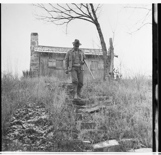 Black and white photograph of Aldo Leopold carrying shotgun down a scrubby hill with small building in background. 